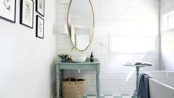 A bathroom with a checkered floor, a white bathtub, a round mirror, a sink with a blue countertop, and framed botanical prints on the wall.