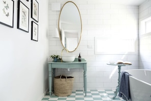 A bathroom with a checkered floor, a white bathtub, a round mirror, a sink with a blue countertop, and framed botanical prints on the wall.