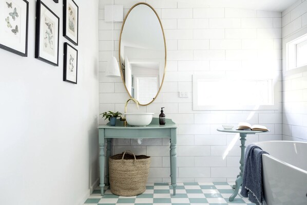 A bathroom with a checkered floor, a white bathtub, a round mirror, a sink with a blue countertop, and framed botanical prints on the wall.