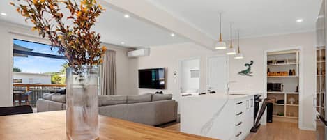 Una cocina moderna con una isla central, un comedor con mesa de madera, un sofá gris y un ventanal grande.