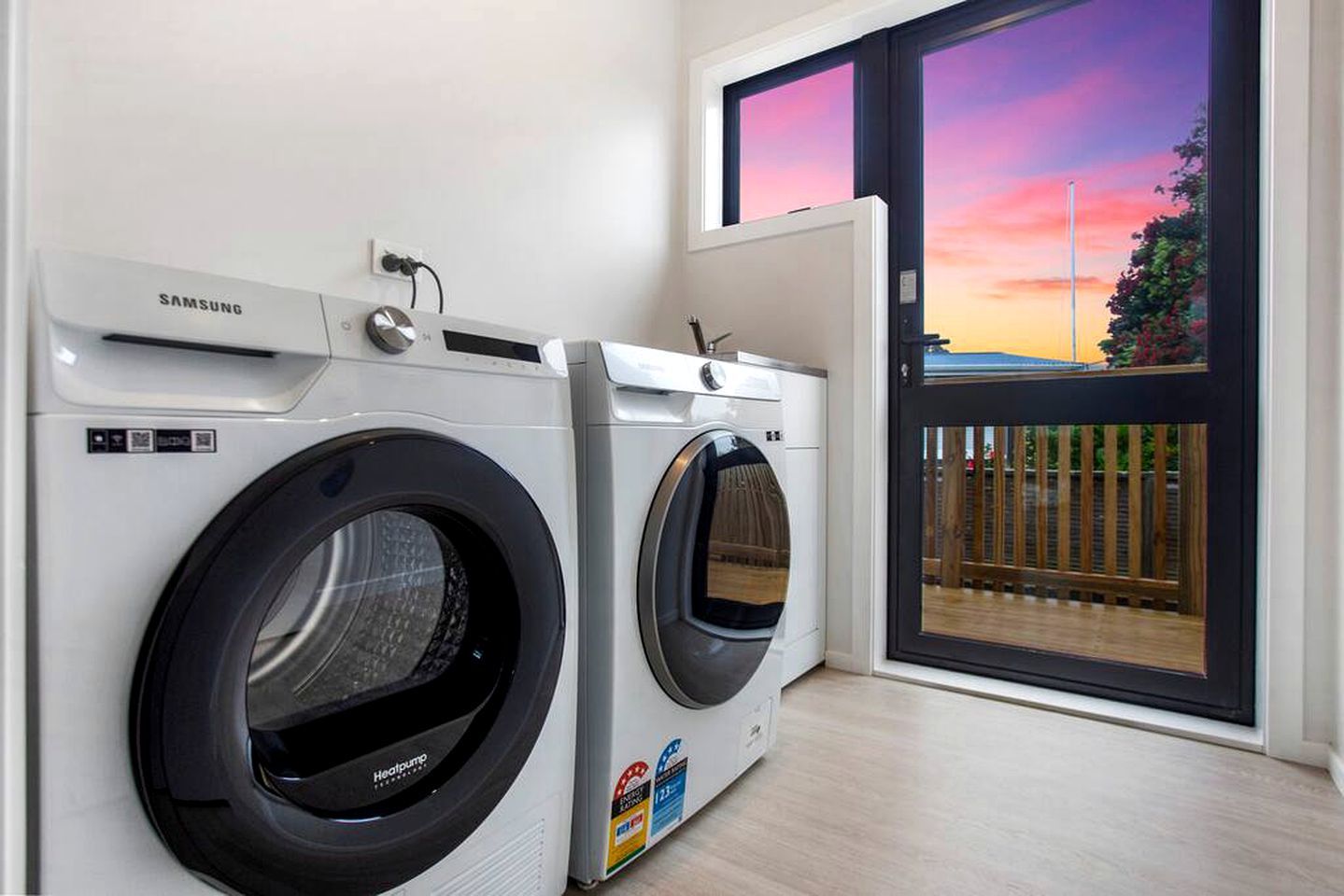 Een wasserette met twee Samsung wasmachines, een raam met uitzicht op een zonsondergang en een deur naar een buitenruimte.