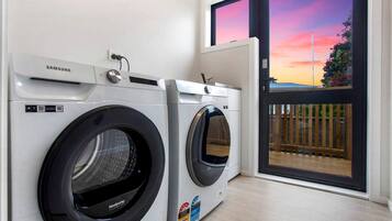 Een wasserette met twee Samsung wasmachines, een raam met uitzicht op een zonsondergang en een deur naar een buitenruimte.