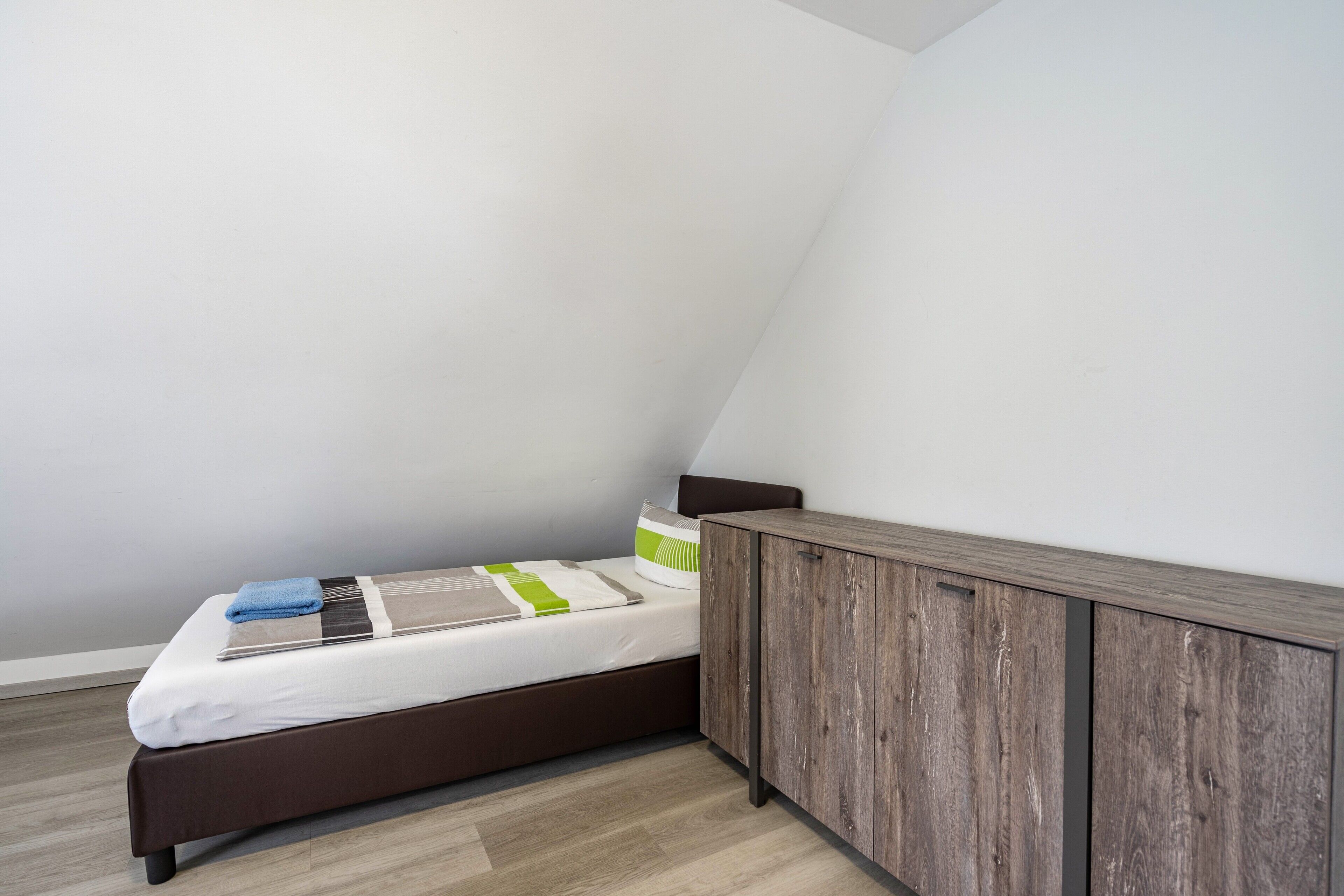 Ein kleines, ordentlich bezogenes Bett mit weißer Matratze und grauem Kopfteil, vor einer weißen Wand mit schrägem Dach.