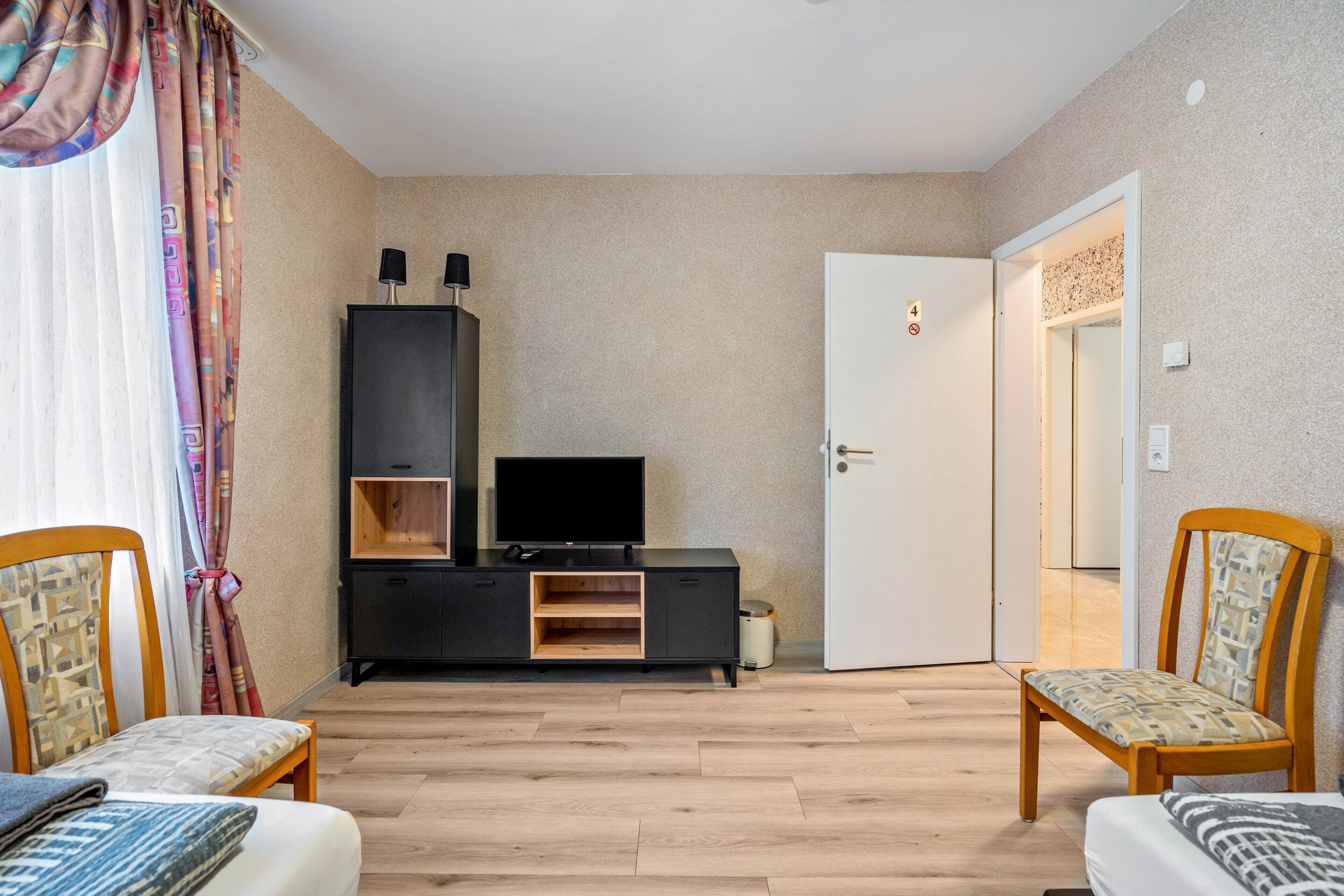 Ein Zimmer mit einem Bett, einem Fernseher auf einem schwarzen Schrank, einer Tür mit einer Nummer und einem Sessel.