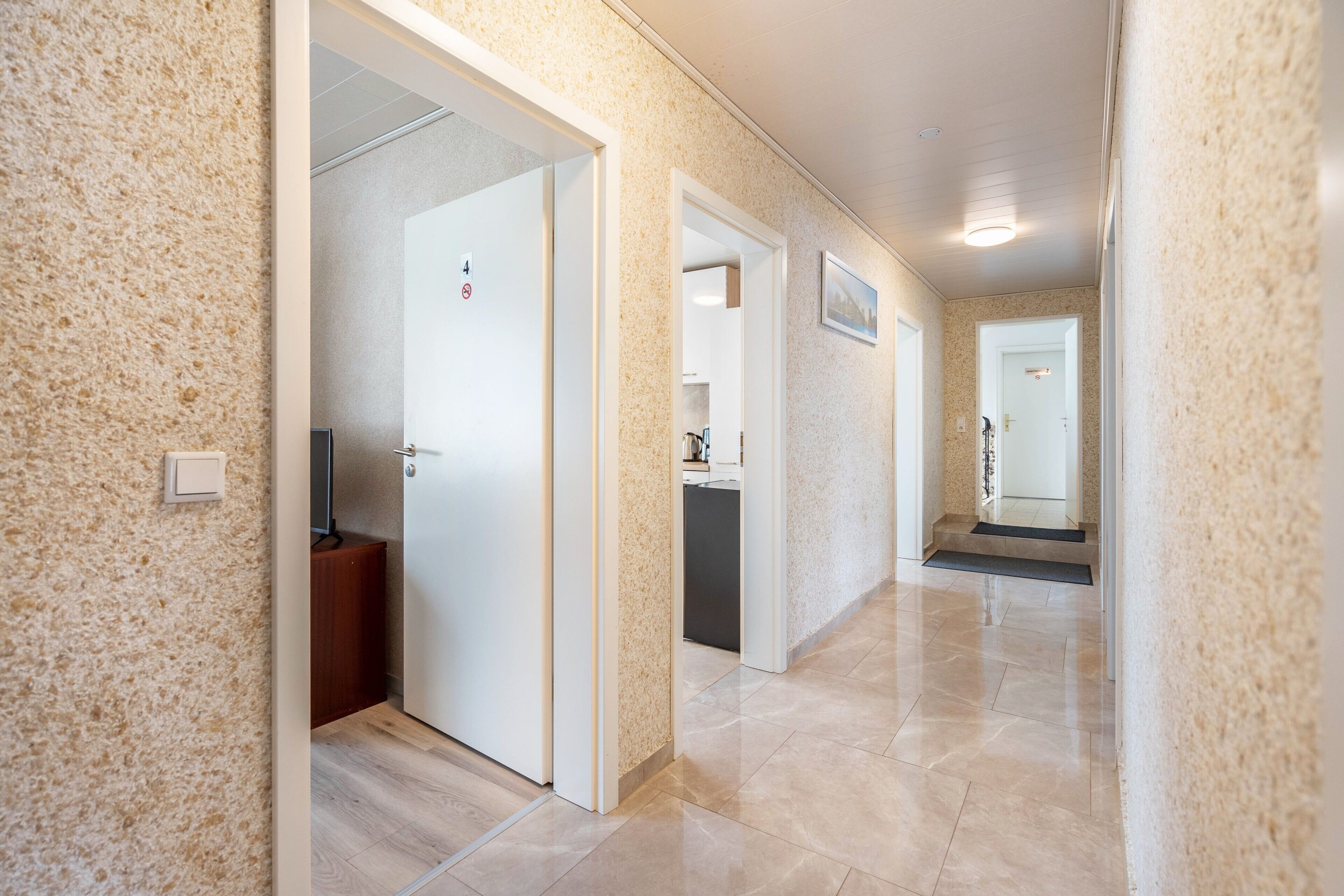 Un couloir doté d’un sol carrelé, de portes blanches et d’un petit coin cuisine visible à travers une porte ouverte.