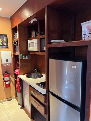 A compact kitchen with a stainless steel refrigerator, a microwave, and a sink. There is a fire extinguisher mounted on the wall, and a shelf with various kitchen items.