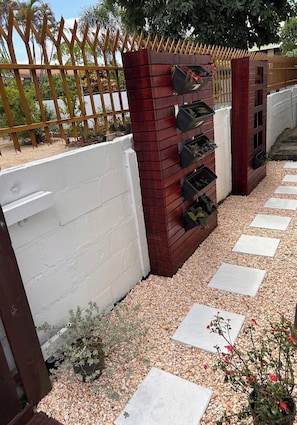 A garden with a white wall, a brick planter with plants, a wooden fence, and stepping stones.