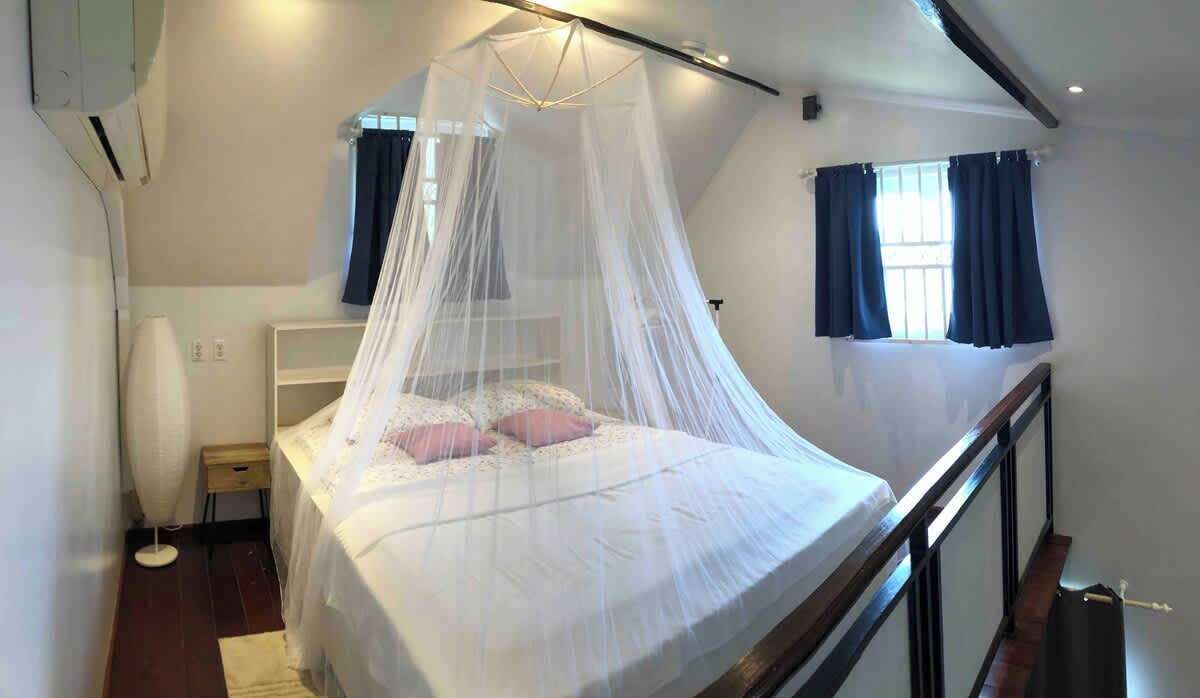 A bedroom with a bed under a canopy, a bedside table, a lamp, and a window with curtains.