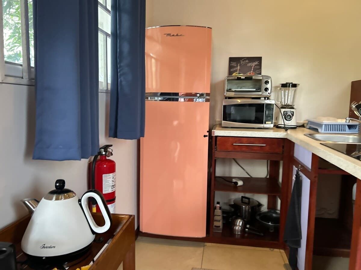 A compact kitchen with a pink refrigerator, a kettle, a toaster, and a fire extinguisher.