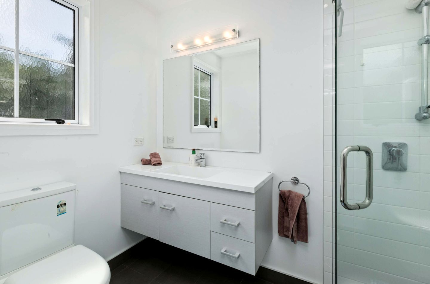A modern bathroom with a white vanity, a large mirror, a glass shower enclosure, and a window.