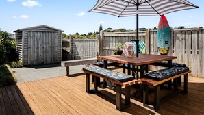 Una terraza de madera con una mesa de picnic y bancos, un paraguas a rayas y una tabla de surf.
