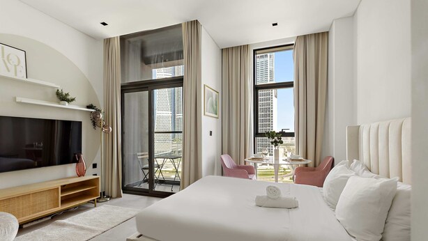 A modern hotel room with a large bed, a flat-screen TV, a balcony with a table and chairs, and a city view. - Luxury Studio in Business Bay With Pool and gym (Dubai)