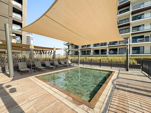 Ein Pool auf dem Dach mit Liegestühlen und Sonnensegel, umgeben von einem modernen Gebäude mit Balkonen.