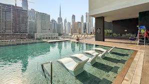A rooftop pool with clear water, lounge chairs, and a view of a city skyline with high-rise buildings. - Luxury Studio in Business Bay With Pool and gym (Dubai)