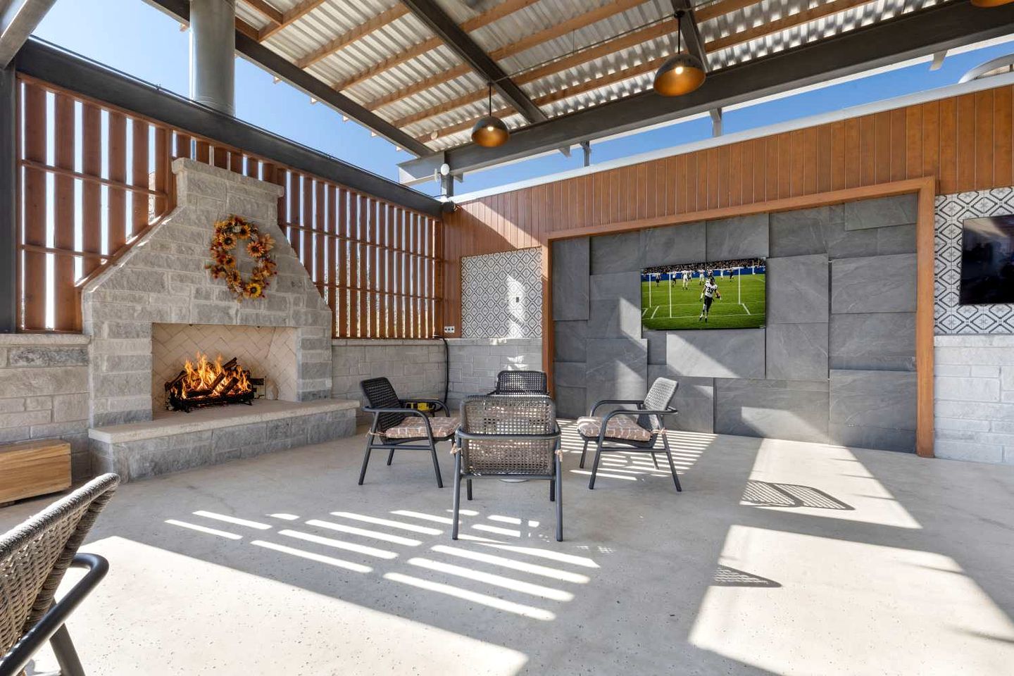 Patio esterno con camino in pietra, mobili in vimini e uno schermo televisivo che mostra una partita sportiva.
