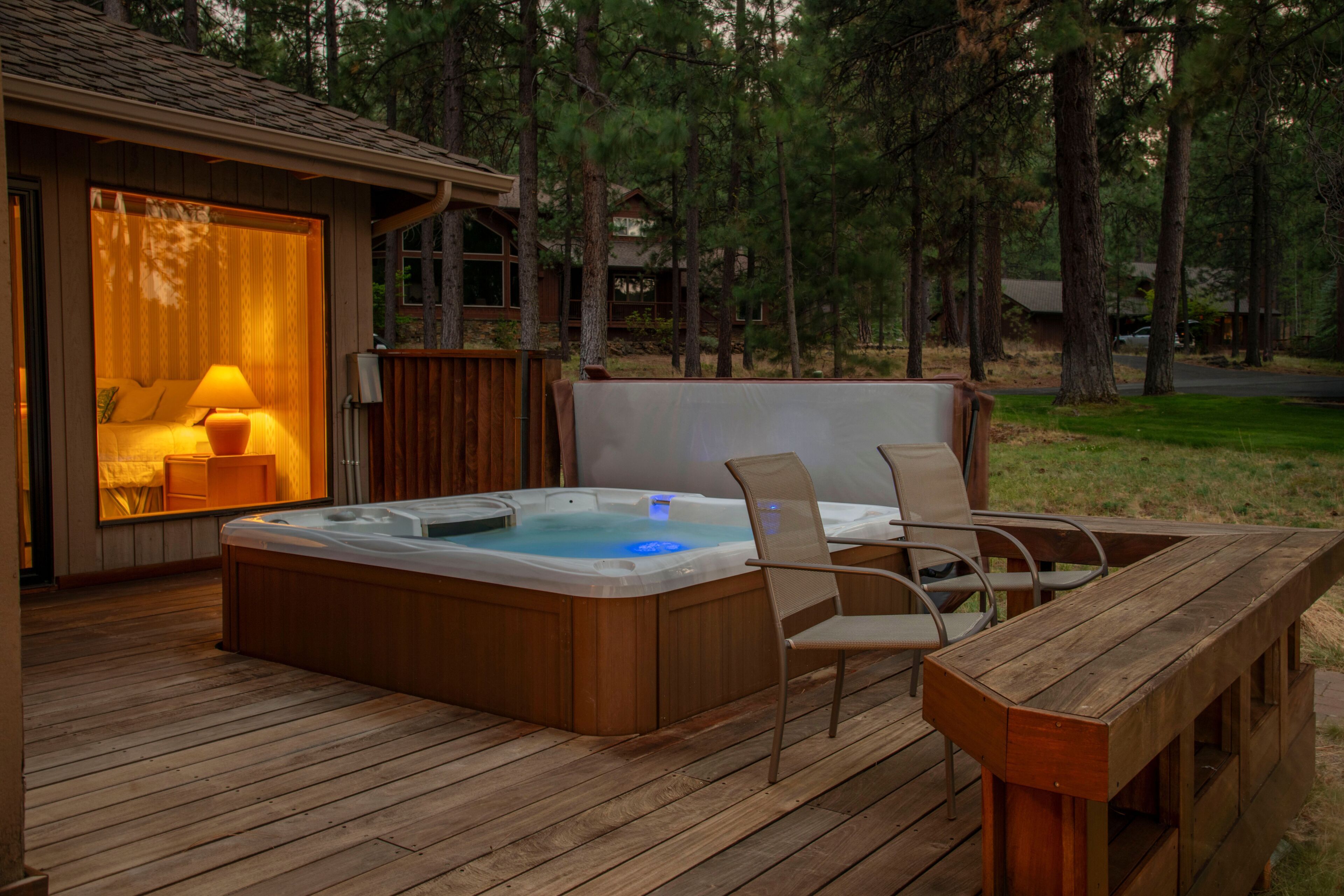 A hot tub on a wooden deck with chairs, a table, and a room with a lamp.