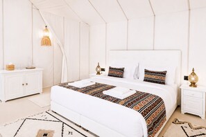 A bedroom with a large bed, white walls, a white wardrobe, and a bedside table with a lamp.