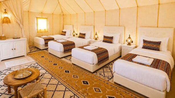 A tent-style room with four beds, a wooden table with glasses, a woven chair, and a patterned rug.