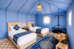 A room with two beds, a hanging light fixture, a small table with a bottle, and a hanging rack.