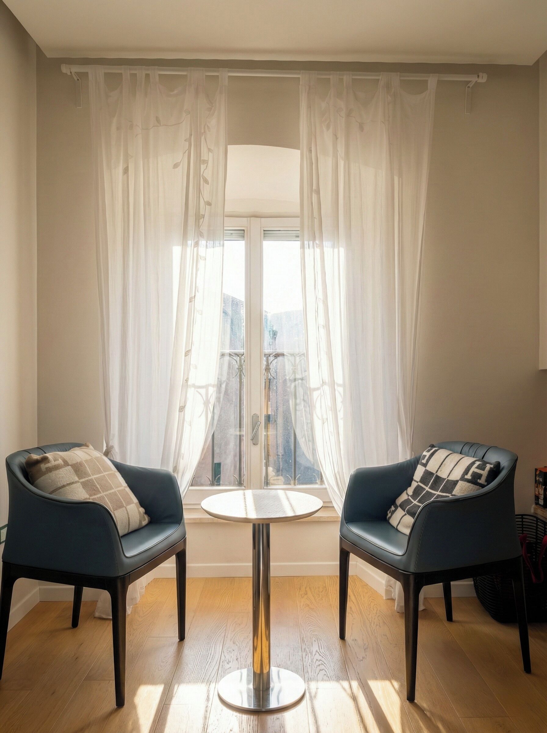 Ein Zimmer mit zwei Sesseln, einem kleinen runden Tisch und Blick durch ein Fenster mit durchsichtigen Vorhängen.