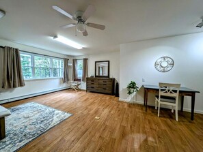 A spacious room with hardwood flooring, a ceiling fan, a dining table with chairs, a dresser, and a large window with curtains. - Serene Lakeside Cabin with Fire Pit, Waterfront Retreat Near Graham Lake, Mariaville Maine (Mariaville)
