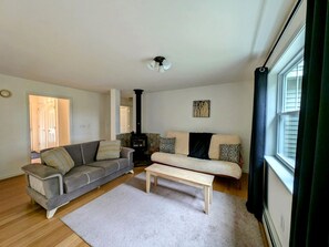Una sala de estar con un sofá gris, un sofá blanco, una mesa de centro de madera, una chimenea y una ventana con cortinas.