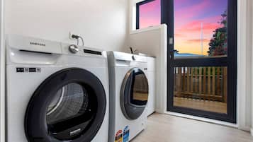 A laundry room with two Samsung washing machines, a window with a view of a sunset, and a door leading to an outdoor area.