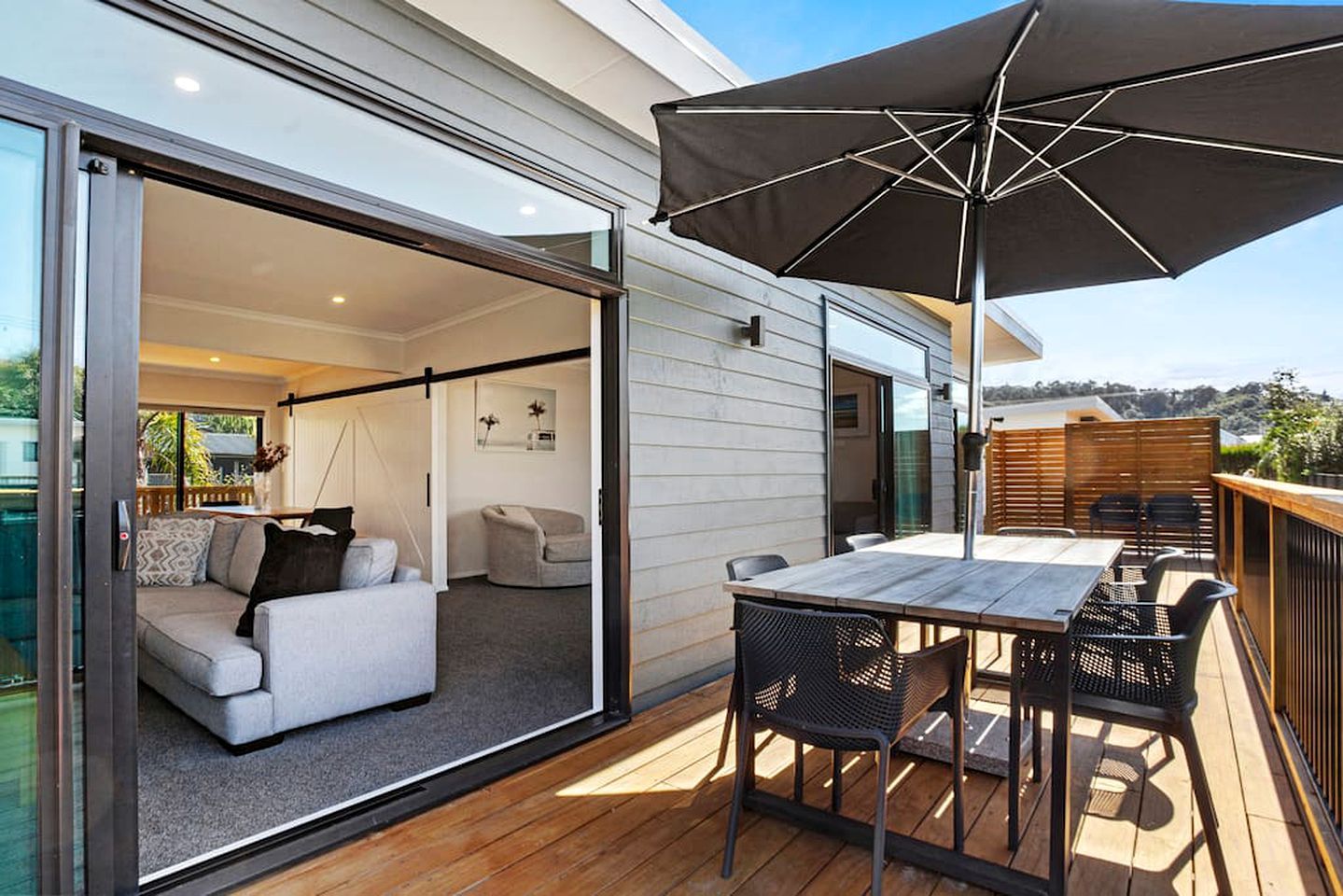 Eine moderne Terrassenanlage mit Tisch und Stühlen, einem großen Sonnenschirm und einer Glasschiebetür zu einem Innenwohnraum.