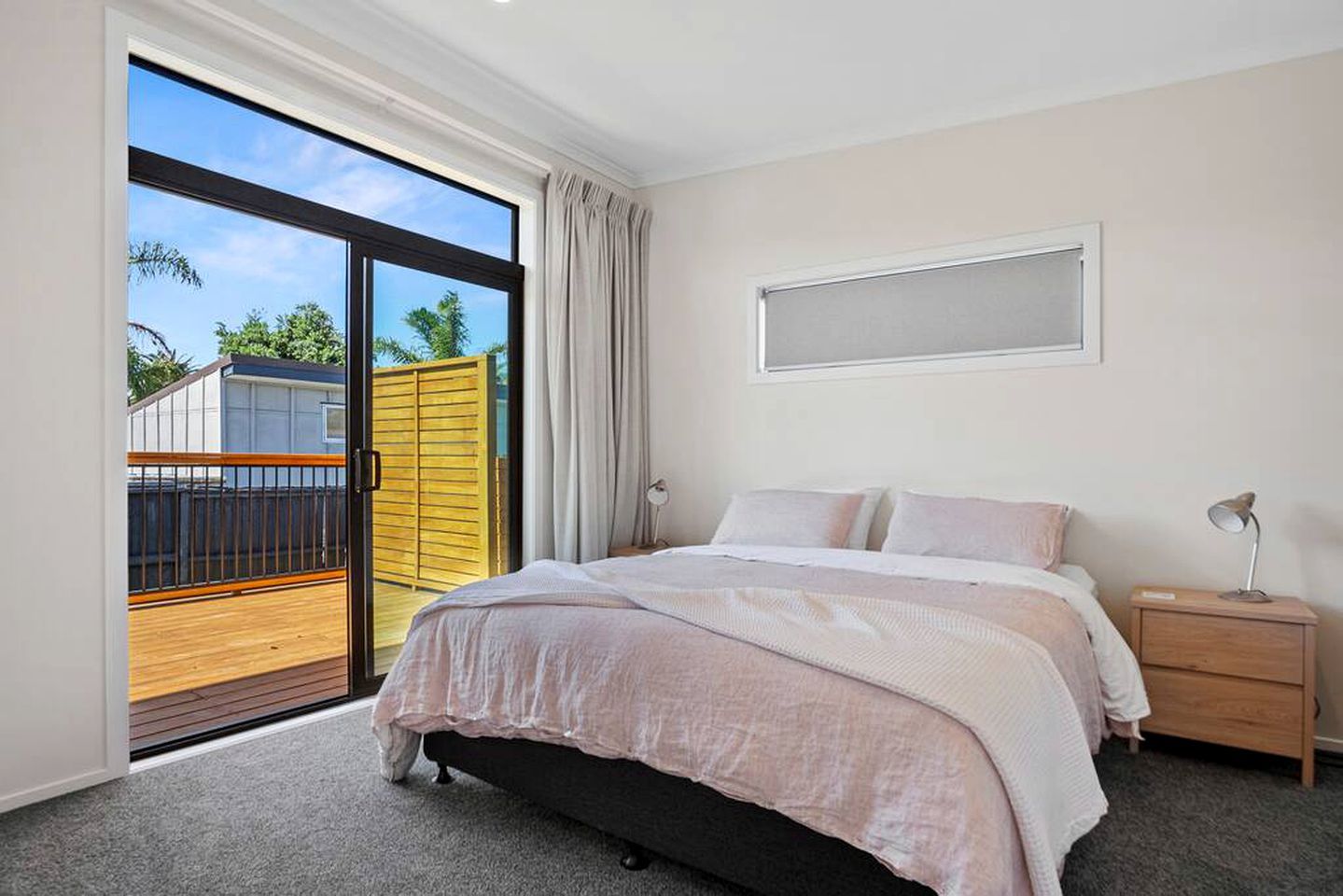 A bedroom with a bed, a nightstand, a lamp, a window, and a view of a balcony.