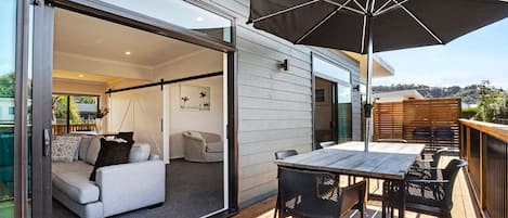 Um deck moderno ao ar livre com mesa e cadeiras, um grande guarda-sol e uma porta de vidro deslizante que dá para um espaço de estar interno.