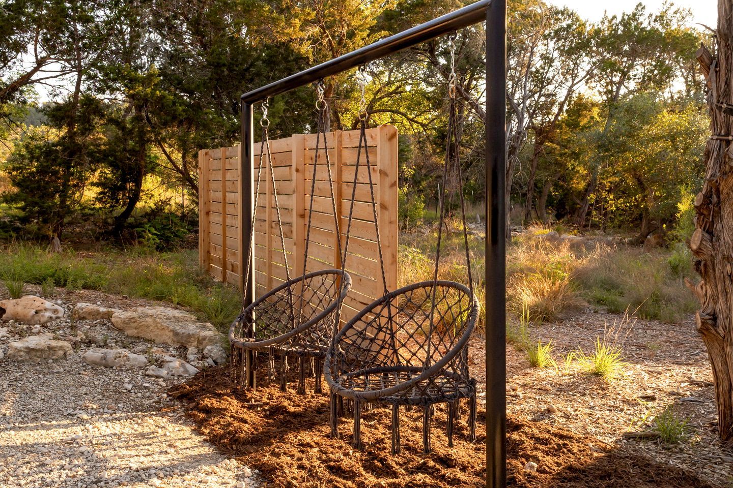 A wooden swing set with two swings, surrounded by a natural landscape with trees and rocks.