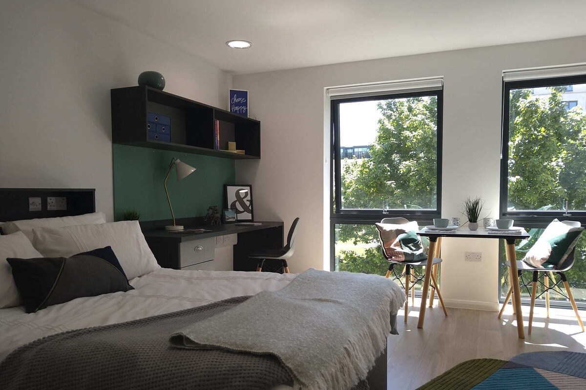A modern bedroom with a bed, desk, chair, and large windows.
