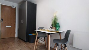 Ein schlichter Raum mit Holztür, dunklem Kleiderschrank, einem kleinen Esstisch mit zwei Stühlen und einer Kakteenpflanze.