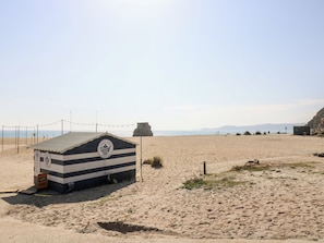 Eine Strandhütte mit gestreiftem Dach und einem Logo, gelegen an einem sandigen Strand mit blauem Himmel und fernen Bergen.