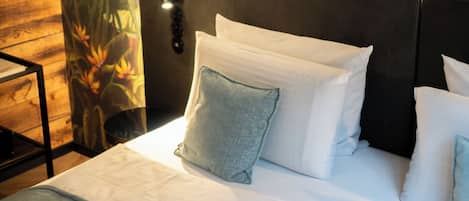 Una cama bien tendida con sábanas blancas y varias almohadas, un cabecero con una lámpara y una pared con un mural tropical.