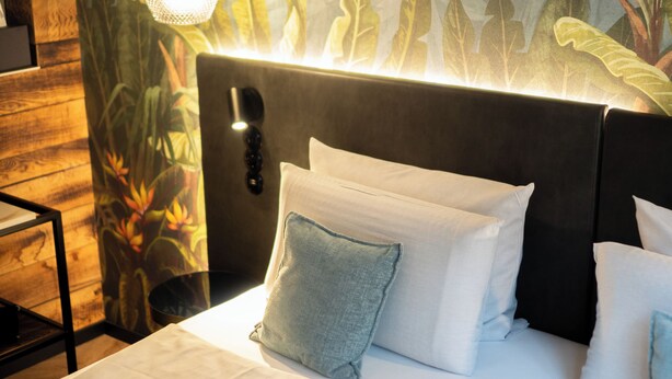 Un lit bien fait, avec du linge de lit blanc et de nombreux oreillers, une tête de lit dotée d’un luminaire et un mur orné d’une fresque aux motifs tropicaux.