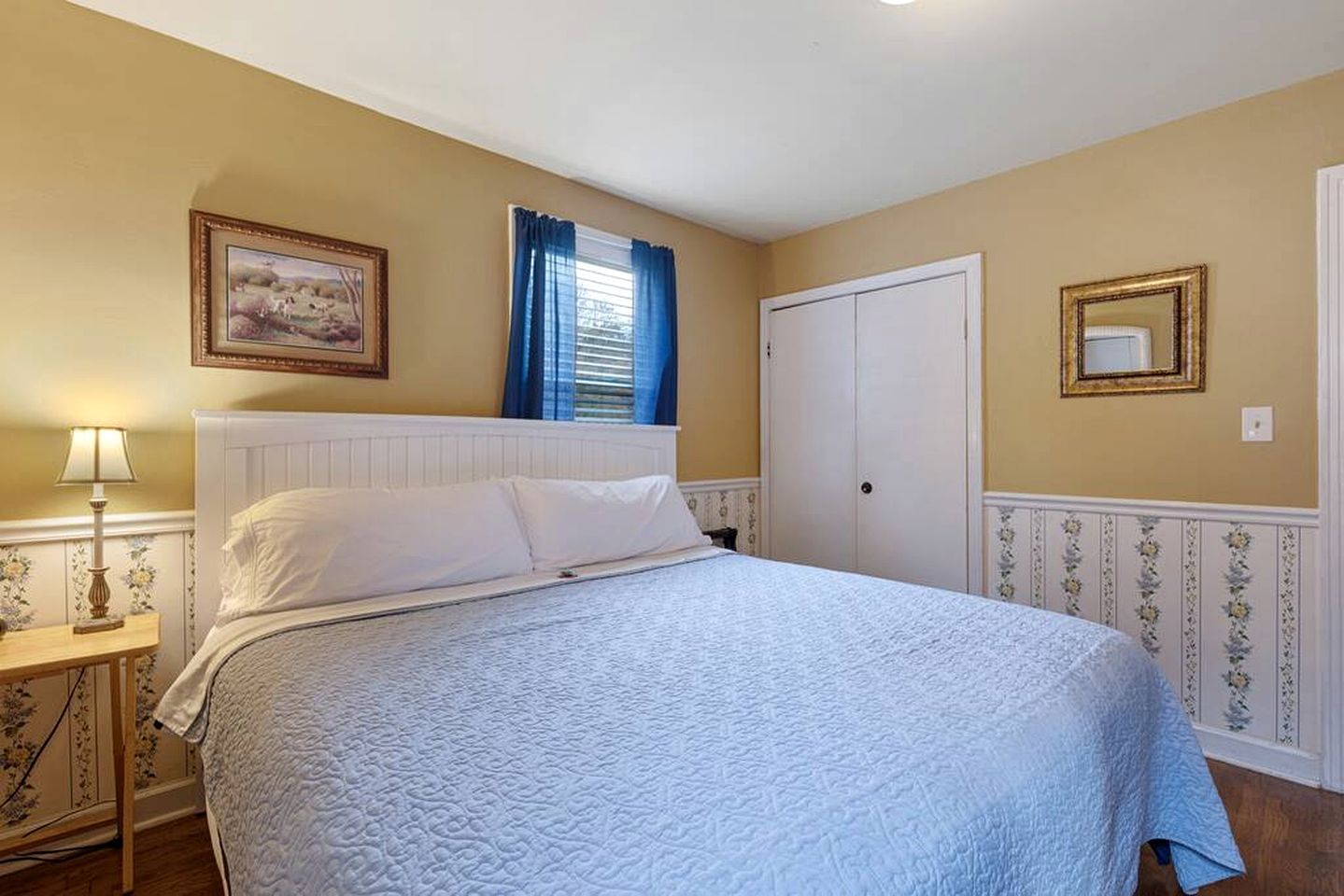 A bedroom with a bed, a nightstand, a lamp, a door, and a framed picture on the wall.
