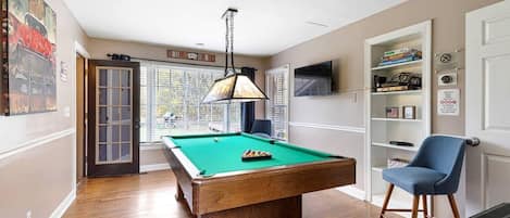 A pool table in a room with a door leading to a balcony, a chair, and a shelf with various items.