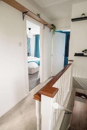 Un pasillo con barandilla de madera, paredes blancas y una puerta que da a un dormitorio con cama y cortinas azules.