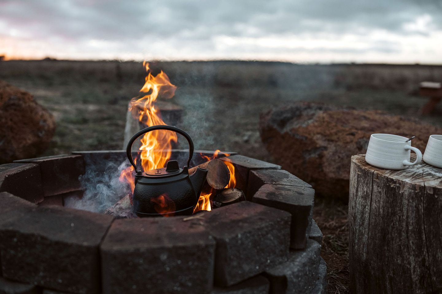A campfire with a kettle and mugs, surrounded by rocks and a wooden stump.