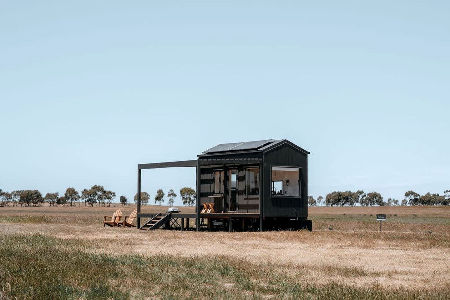 A small, dark-colored cabin with a deck and outdoor seating area, surrounded by a grassy field and trees.