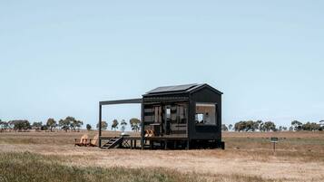 A small, dark-colored cabin with a deck and outdoor seating area, surrounded by a grassy field and trees.