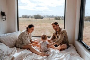 A family of three on a bed with a view of a grassy field through a window.