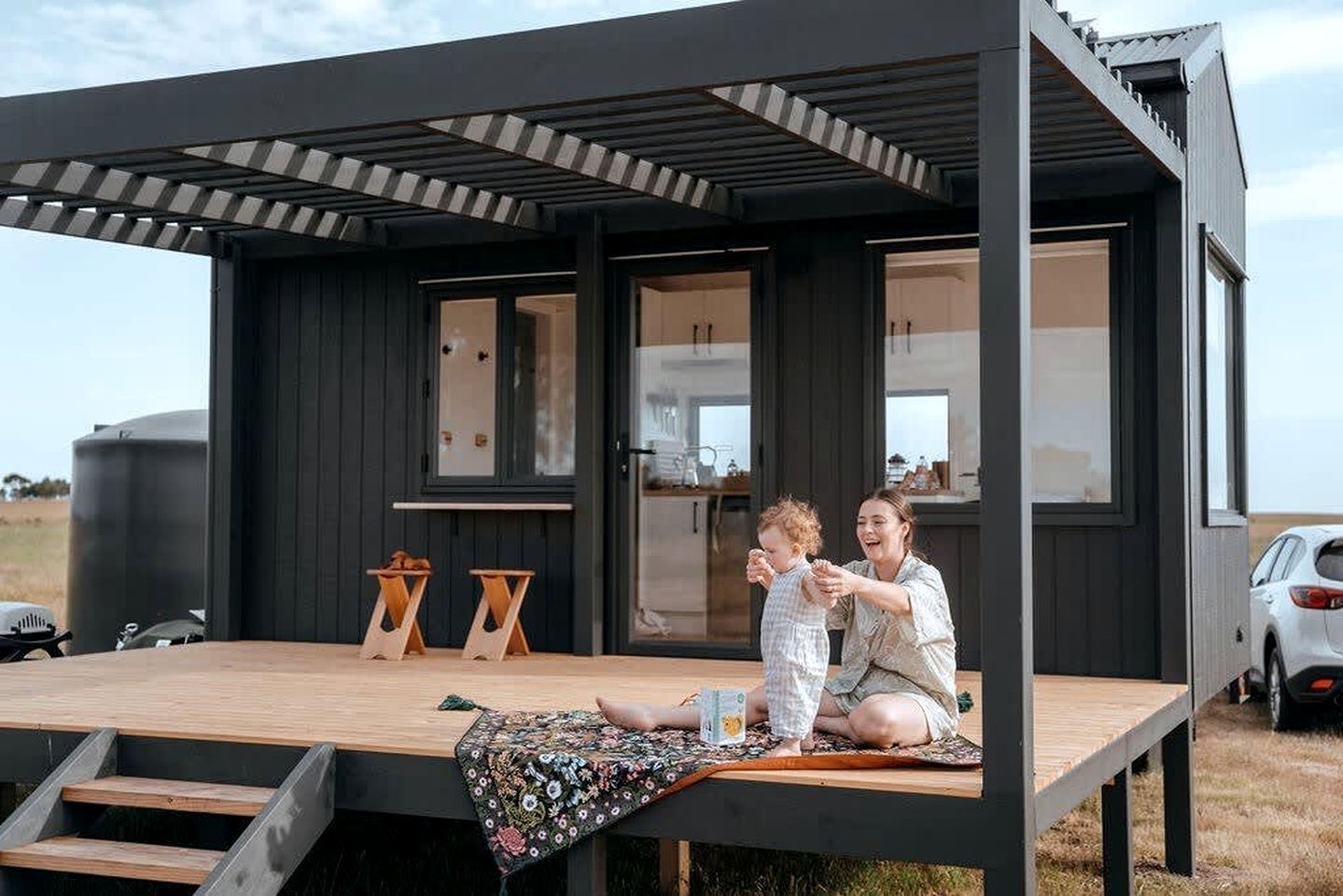A modern, dark-colored cabin with a wooden deck, a small table, and a bench.
