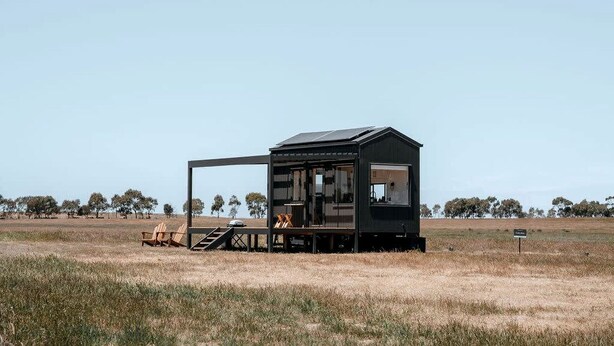 A small, dark-colored cabin with a deck and outdoor seating area, surrounded by a grassy field and trees. - Magical Tiny House with Amazing Stargazing and Deck in Victoria (Skipton)