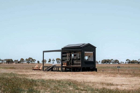 A small, dark-colored cabin with a deck and outdoor seating area, surrounded by a grassy field and trees.