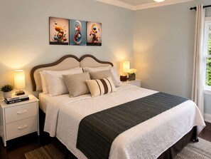 Een netjes opgemaakt bed met een donker hoofdbord, een nachtkastje met een lamp en een muur met drie abstracte schilderijen.