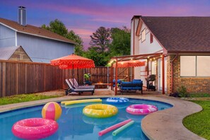 A backyard with a swimming pool, inflatable rings, a covered seating area, and a barbecue grill.