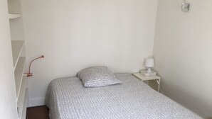 Een kleine, verzorgde slaapkamer met een enkel bed, een nachtkastje met een lamp en een wandlamp.