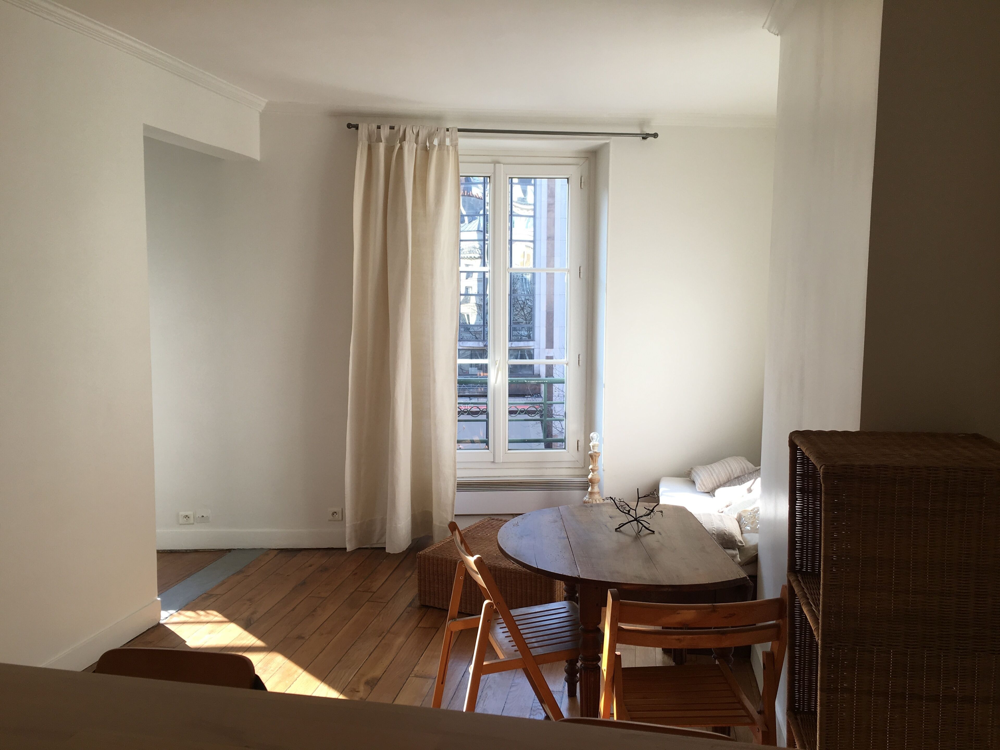 Una habitación con piso de madera, una mesa redonda con sillas, una ventana con cortinas y un banco de mimbre.